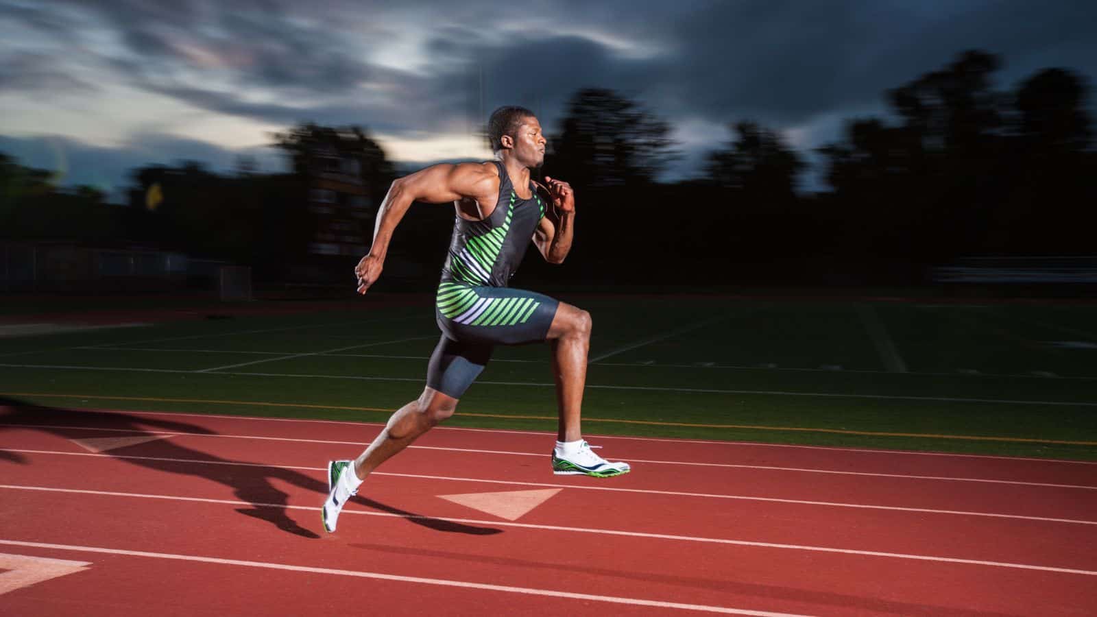 Sprinter mid-stride on track at dusk, wearing black and green suit.